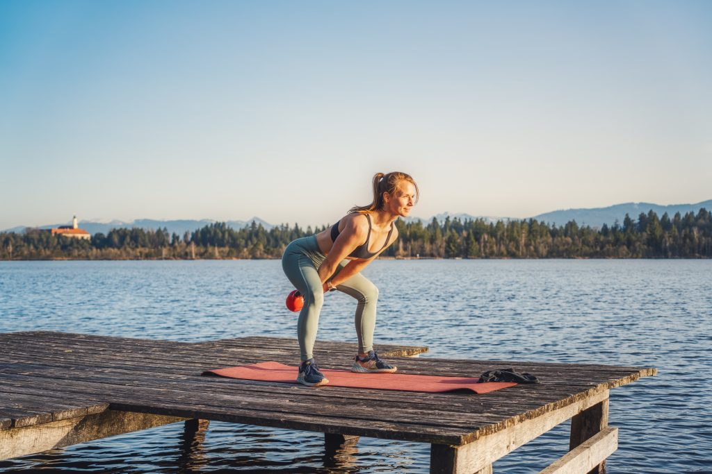 Trainerin Desiree auf einer Trainingsmatte auf einem Steg am See bei der Ausführung einer funktionellen Übung im Stehen, den Kettlebell Swings.