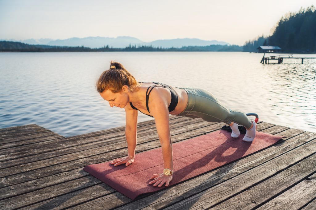 Trainerin Desiree auf einer Trainingsmatte auf einem Steg am See bei der Ausführung einer Bauchübung im hohen Plank.