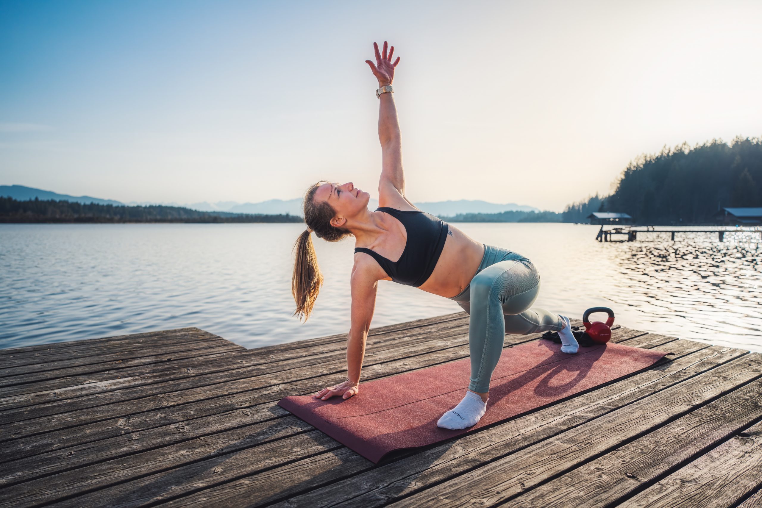 Trainerin Desiree auf einer Trainingsmatte auf einem Steg am See bei der Ausführung einer Mobilitätsübung im tiefen Ausfallschritt zum vorderen Bein aufgedreht, Arm und Blick zeigen zum Himmel, Übung World Greatest Stretch.