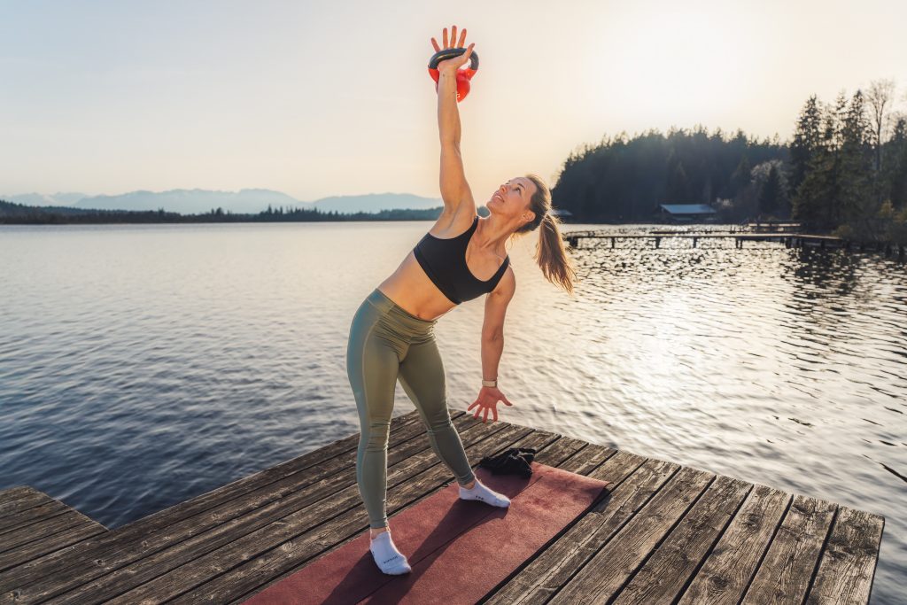 Trainerin Desiree auf einer Trainingsmatte auf einem Steg am See bei der Ausführung einer Rumpfstabilitätsübung im Stehen mit Extragewicht, Kettlebell am gestreckten Arm über Kopf, anderer Arm führt zum Fuß hin, breiter Stand, tiefe Beuge seitlich, bei der Übung Windmill mit Kettlebell.