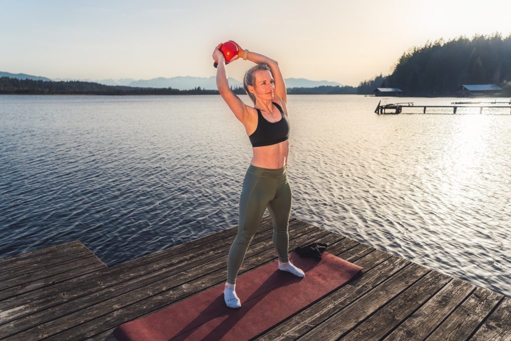 Trainerin Desiree auf einer Trainingsmatte auf einem Steg am See bei der Ausführung einer Bauchübung im Stehen mit Extragewicht, Kettlebell über Kopf um den Körper führend für gesamte Rumpfstabilität.