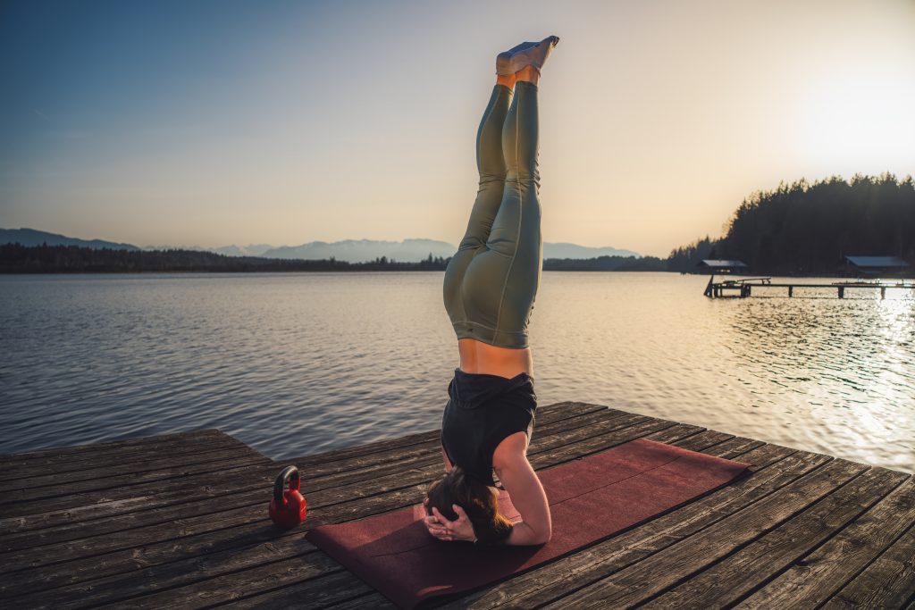 Trainerin Desiree im Kopfstand auf einer Trainingsmatte auf einem Steg am See, entspannt nach dem Training. Neben der roten Matte steht eine rote Kettlebell für funktionelles Training.