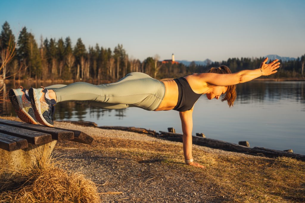 Trainerin Desiree mit Füßen auf der band und Händen auf dem Boden, einen Arm nach vorn gestreckt, einen hohen Plank mit erhöhter Instabilität ausführend.
