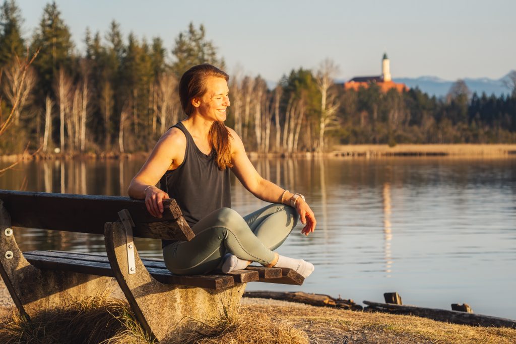 Profilbild der Trainerin Desiree, herzhaft lachend, auf einer Bank am See sitzend im Schneidersitz und Sportkleidung.