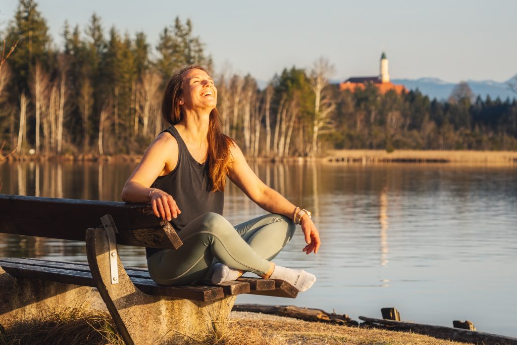 Profilbild der Trainerin Desiree, herzhaft lachend, auf einer Bank am See sitzend im Schneidersitz und Sportkleidung.