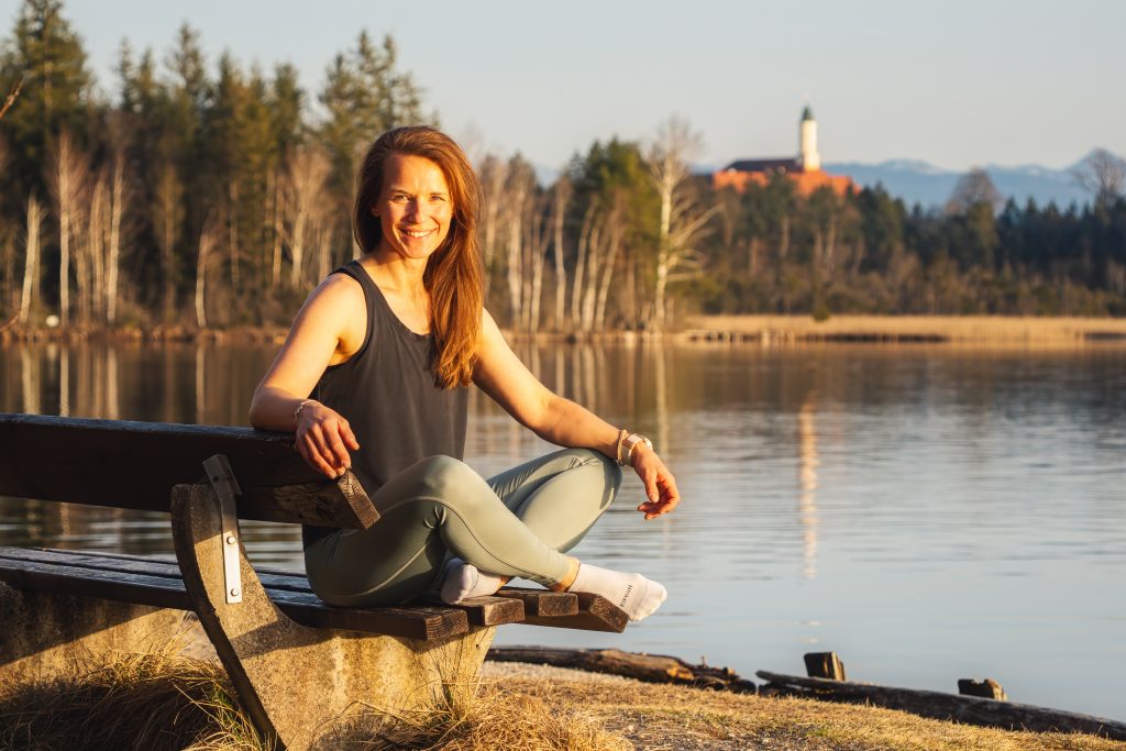 Profilbild der Trainerin Desiree, freundlich in die Kamera lächelnd, auf einer Bank am See sitzend.