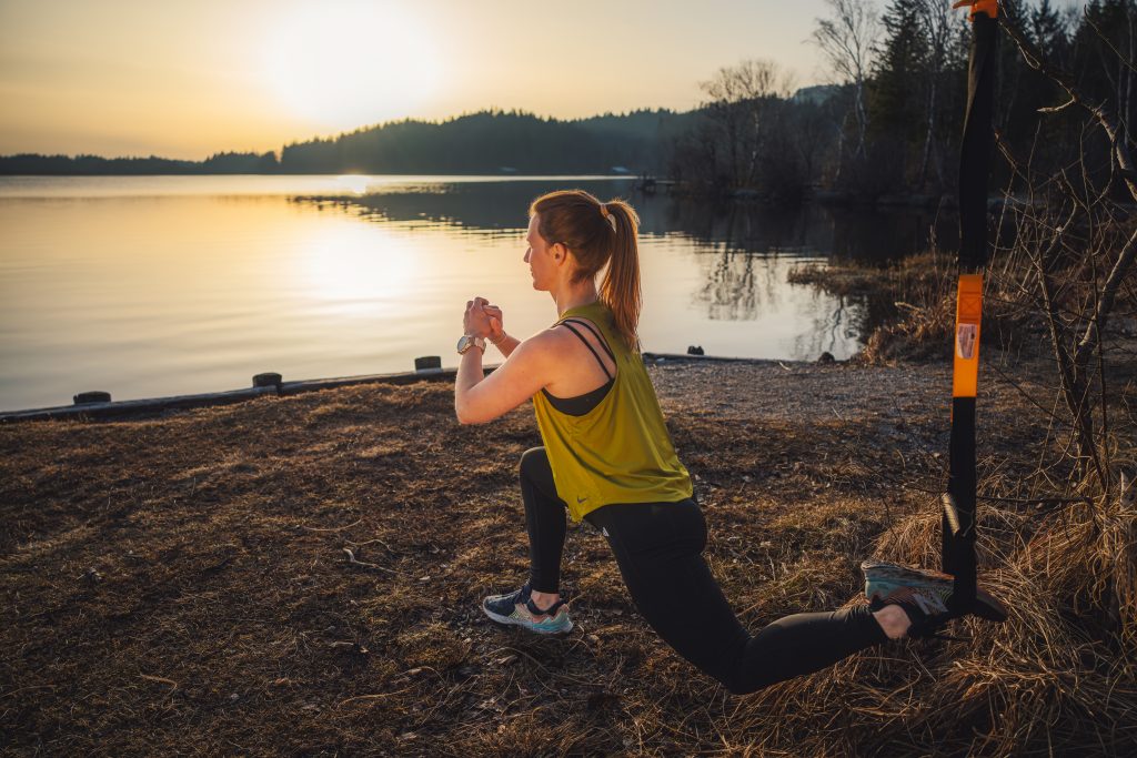 Trainerin Desiree bei der Ausführung einer funktionellen Übung Outdoor. Mit einem gelben TRX Schlingentrainer an einem Baum befestigt, ein Fuß in der Schlaufe, tiefer Ausfallschritt, bei der Übung tiefer reverse Lunge mit hinterem Fuß in der Schlinge.