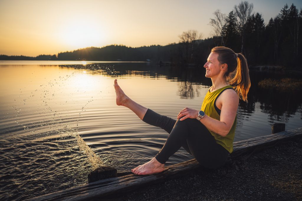 Trainerin Desiree am Ufers eines Sees sitzend und mit Freude mit den Füßen im Wasser spielend.