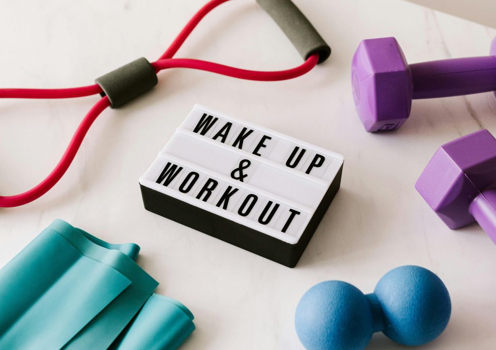Aufforderung zu "Wake up & Work out" mit motivierenden Kleingeräten wie Hanteln und Gummiband um das Schild herum für ein funktionelles Home Workout